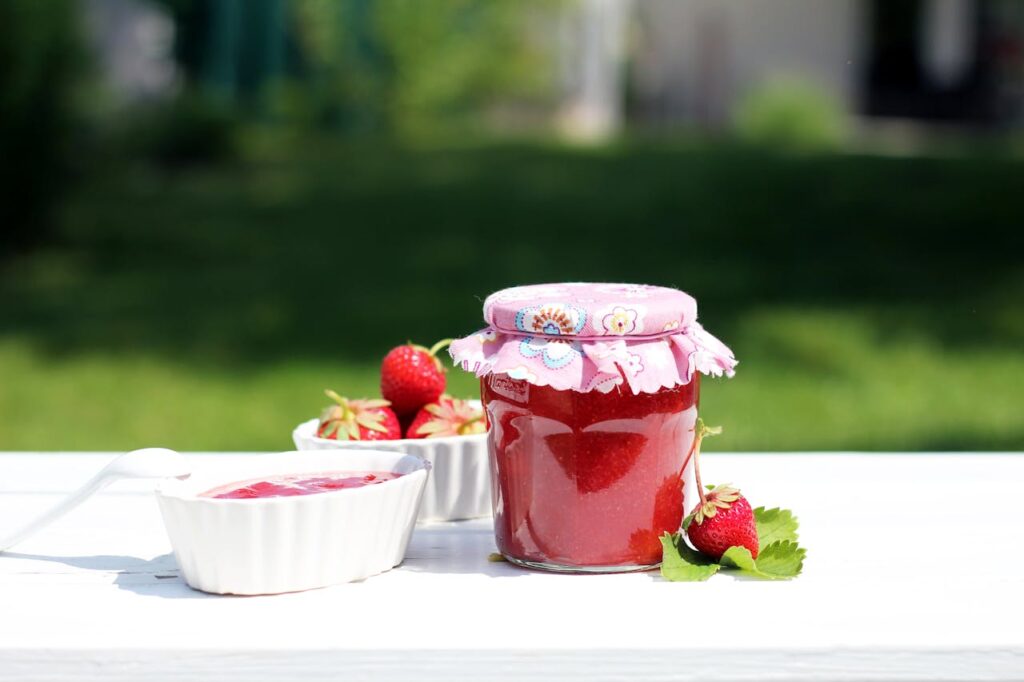 Fresh homemade strawberry jam in a decorative jar with ceramic bowls on a white table outdoors.